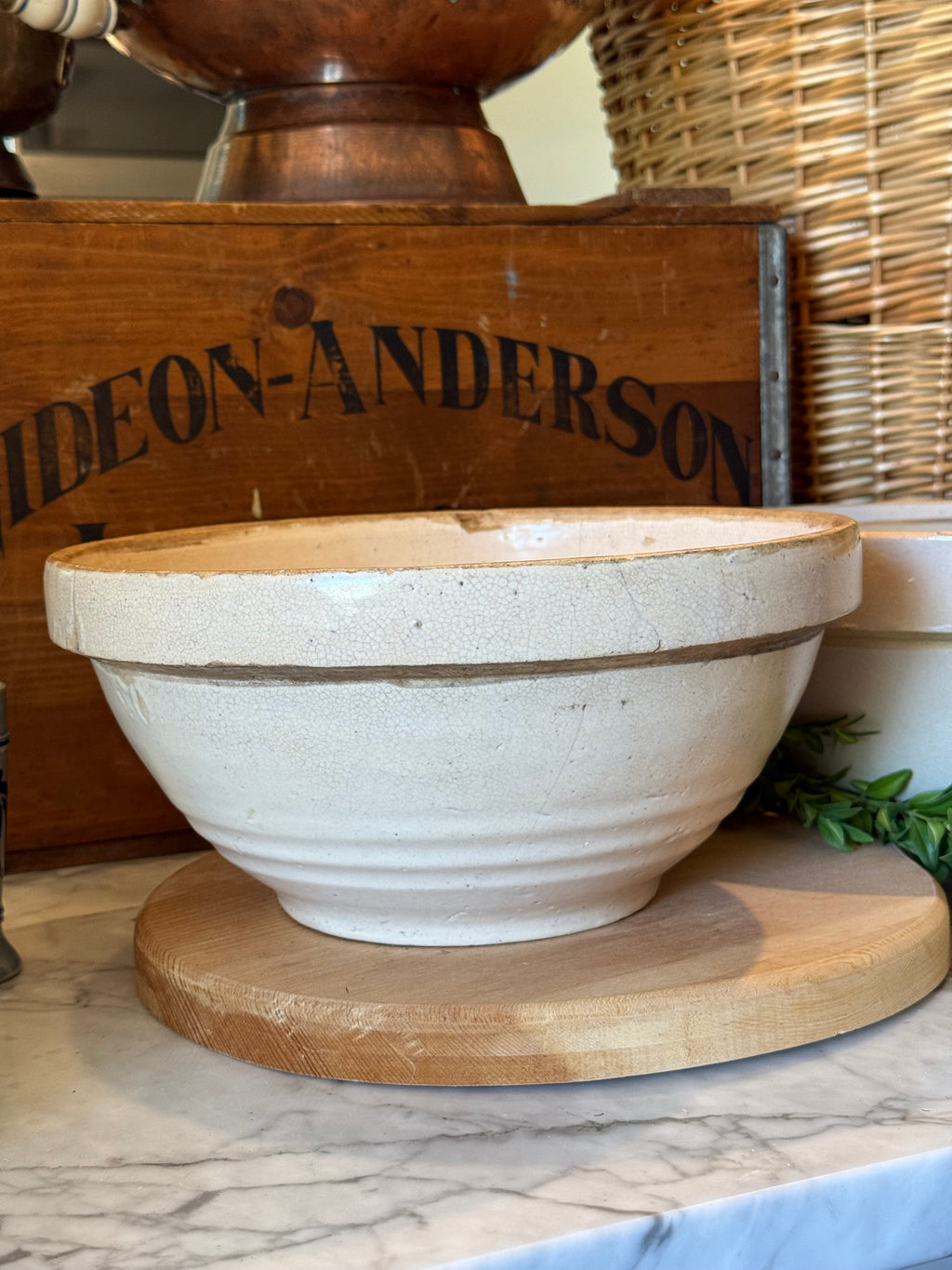 A large off-white ceramic mixing bowl with a rustic finish, displayed on a round wooden board on a marble countertop. In the background, a wooden crate visible next to a woven basket.