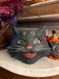 A black cat-shaped decorative mask with green eyes, white whiskers, and an orange nose, displayed indoors next to a white curved surface and purple leaves.
