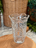 A clear cut-crystal vase with a leaf pattern and scalloped rim, displayed on a wooden surface with a wicker basket and green plants in the background.