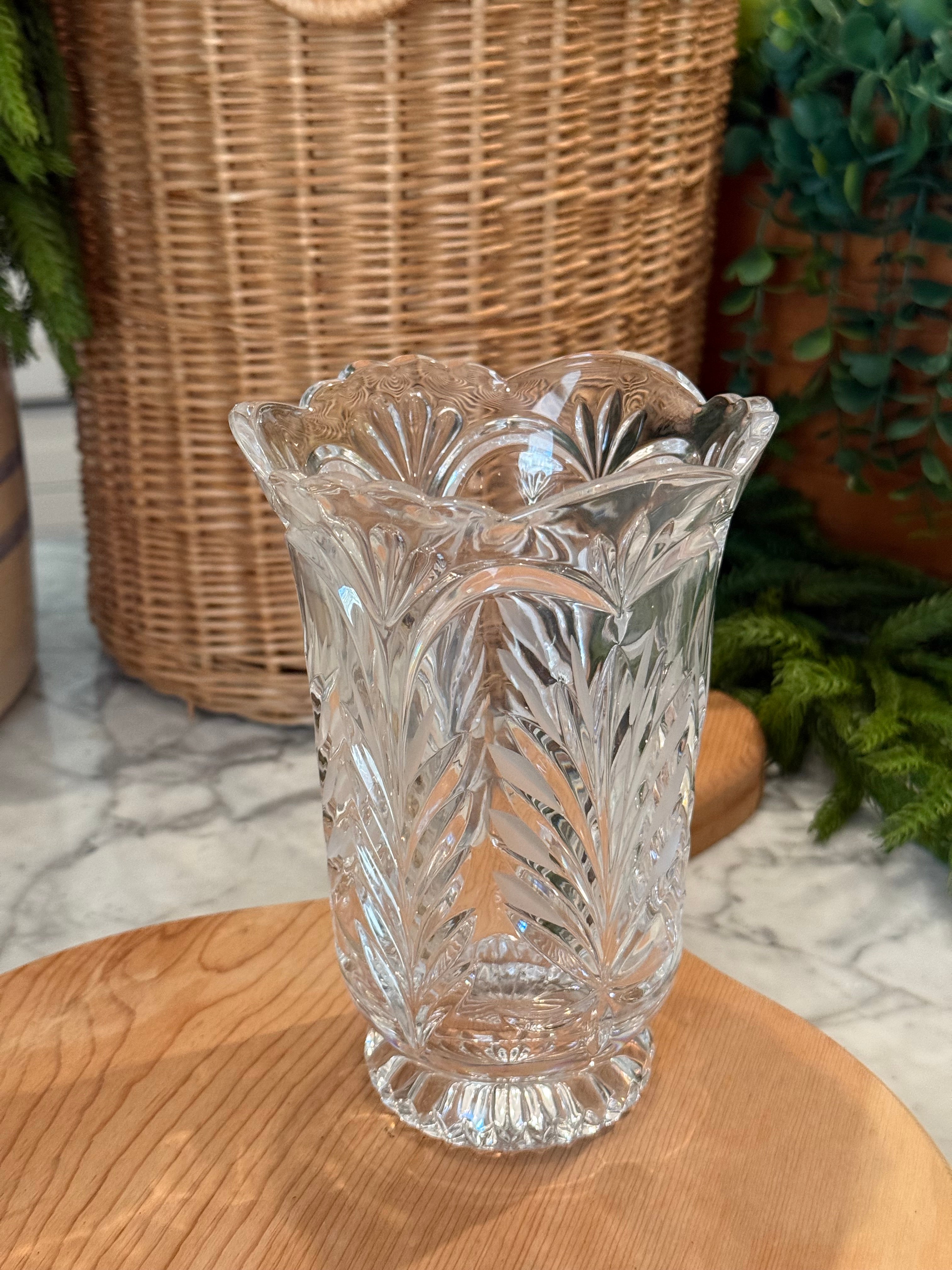 A clear cut-crystal vase with a leaf pattern and scalloped rim, displayed on a wooden surface with a wicker basket and green plants in the background.