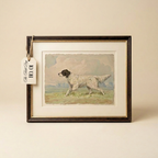 A framed vintage-style print showing a white and black dog running across a grassy field under a cloudy sky, mounted with a wide cream mat and a dark wood frame. A small hanging tag is attached to the left side.