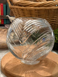 A clear glass decorative bowl with etched wavy leaf-like patterns, displayed on a round wooden stand with a wicker basket and books in the background.