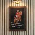 Archival 1909 equestrian print of jumping horse framed in warm walnut burl wood, hanging in a cozy traditional study with a collegiate, quiet luxury aesthetic.