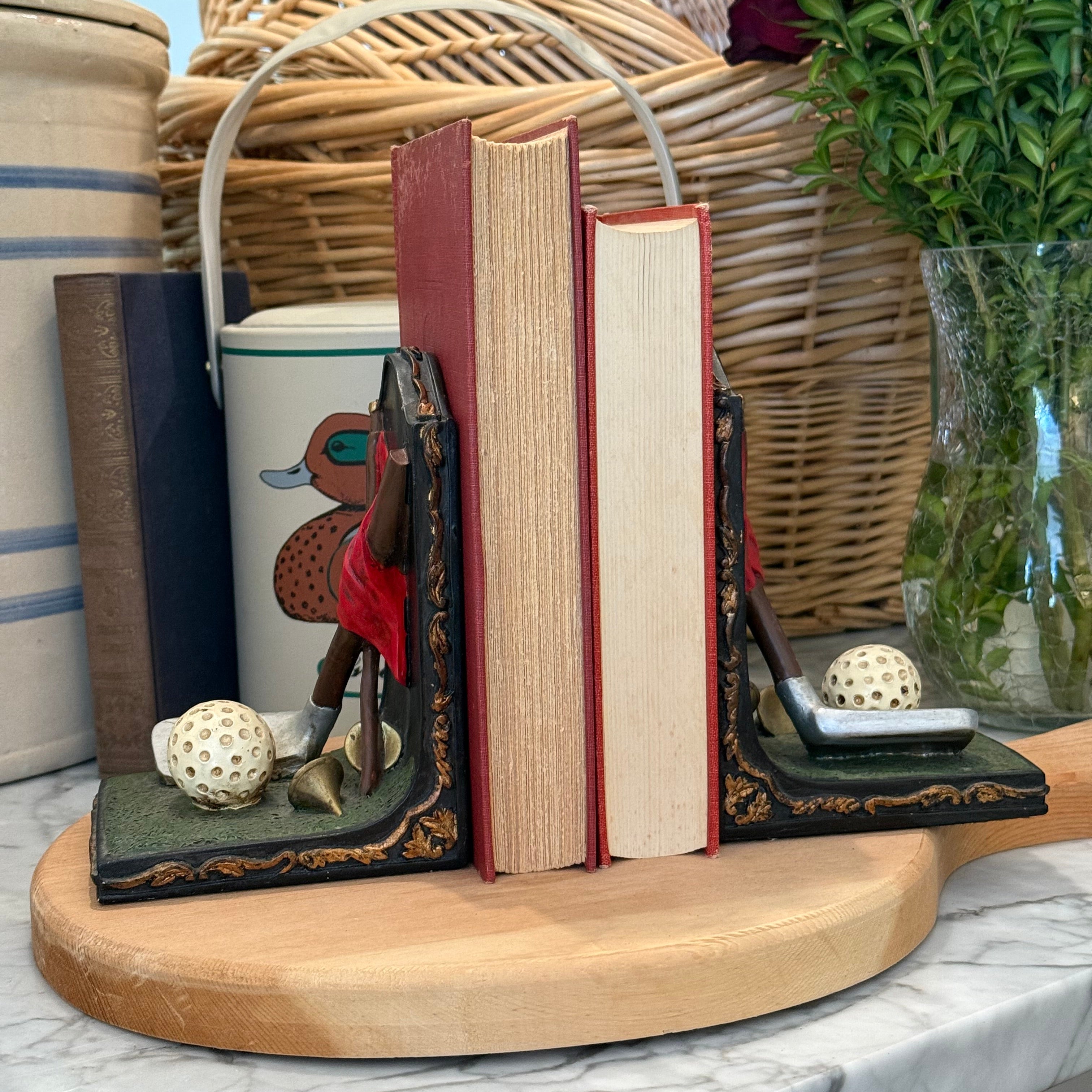 Decorative bookends holding two vintage-style books on a round wooden tray, styled on a marble surface with a wicker basket, ceramic mugs, and a glass vase of flowers in the background.