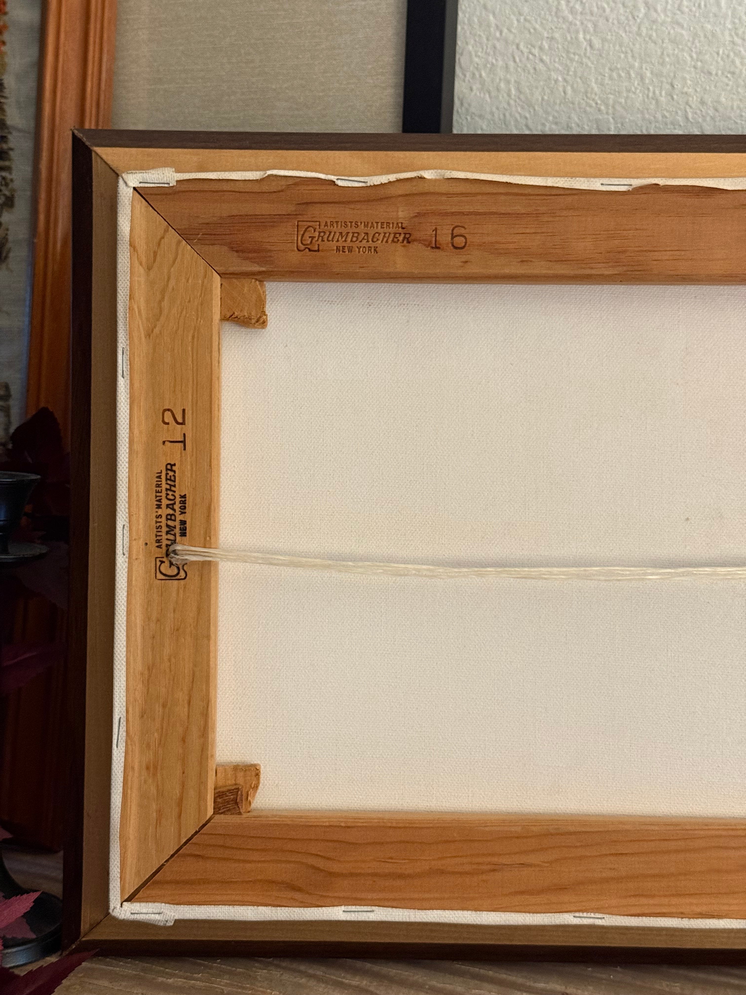Close-up of the back of a framed artwork or canvas, showing a light wood frame, off-white canvas surface, hanging cord, and printed maker marks near the bottom.