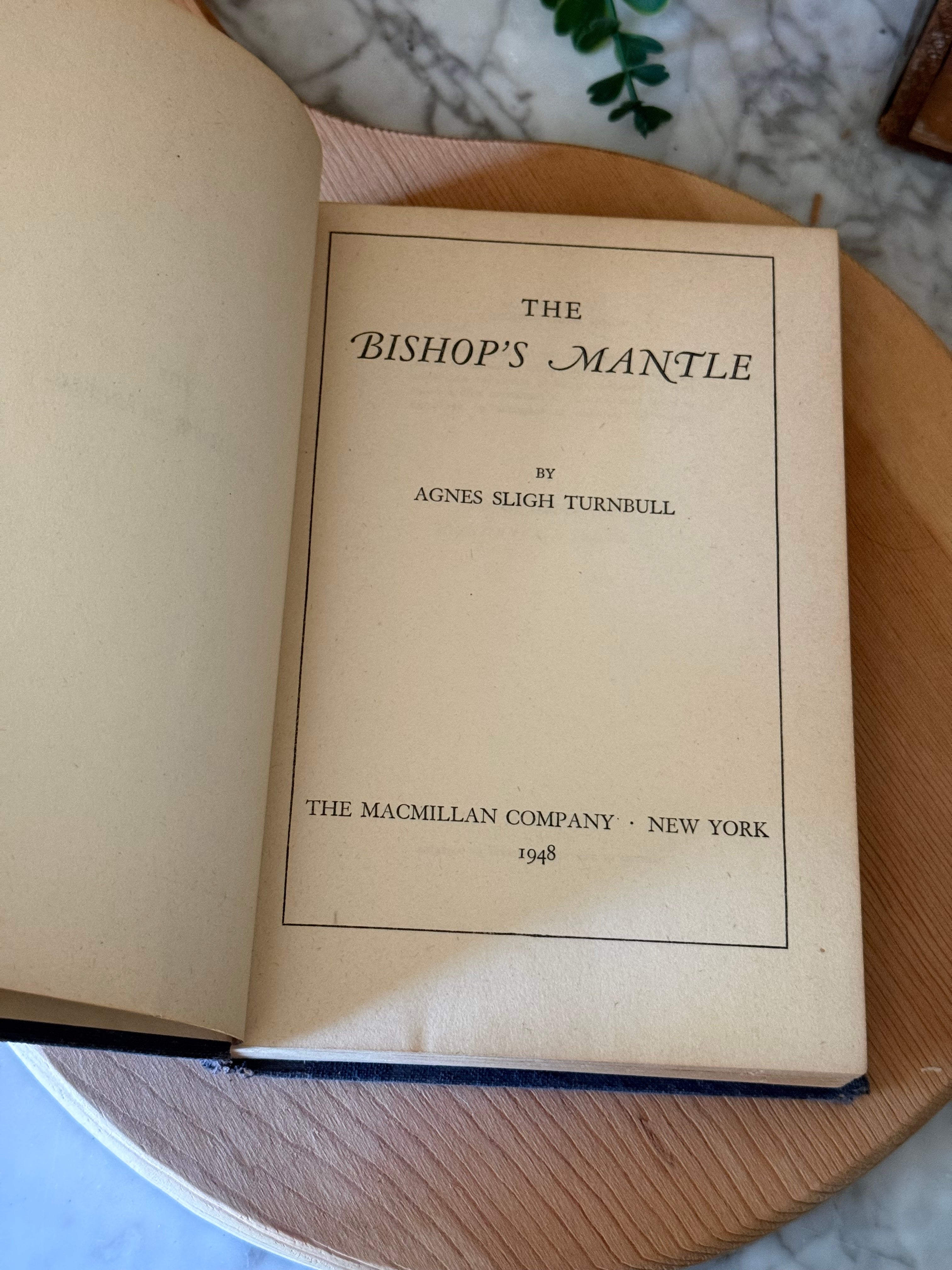 An open vintage book titled 'The Bishop's Manacle' by Agnes Smith Turnbull, showing the title page with aged cream paper, placed on a round wooden tray over a marble surface.