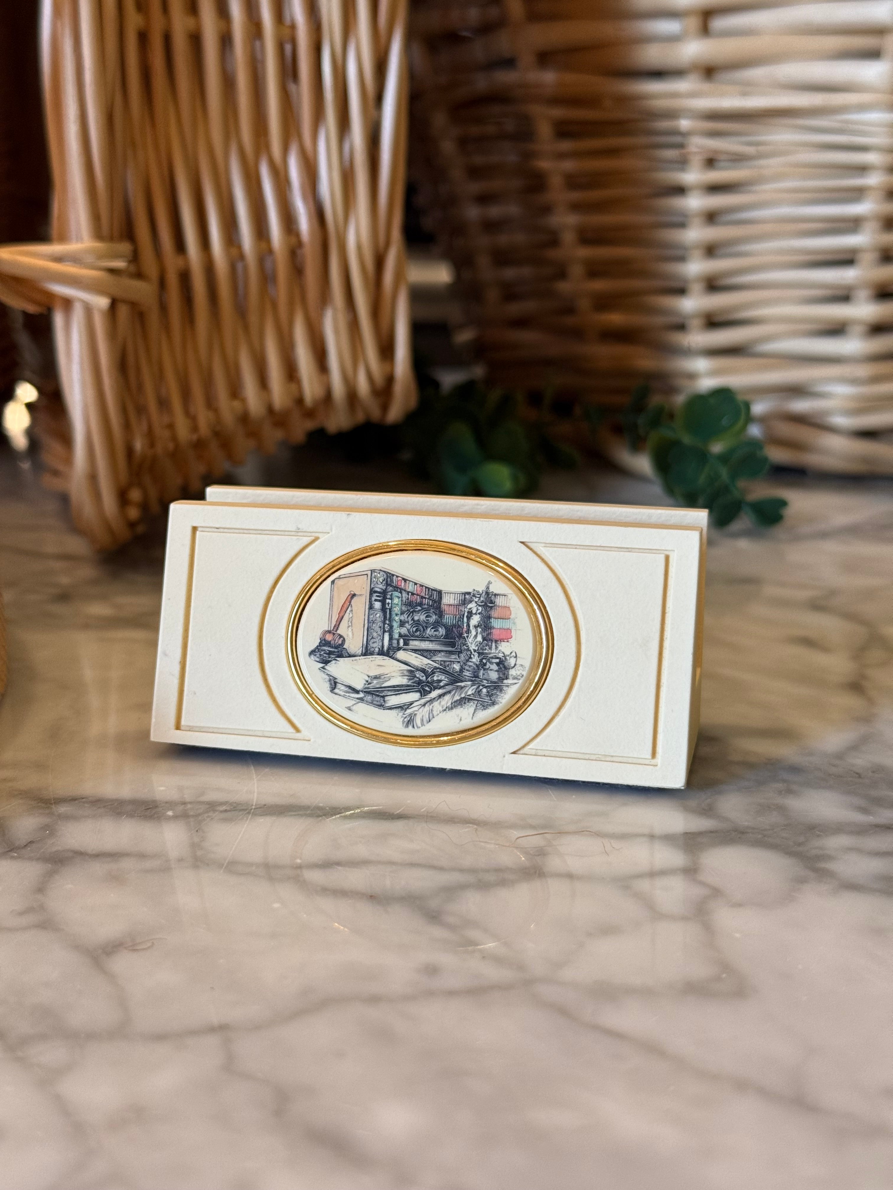 A small rectangular cream-colored decorative box or card holder with gold trim and an oval illustration on the front, placed on a marble surface with wicker baskets in the background.