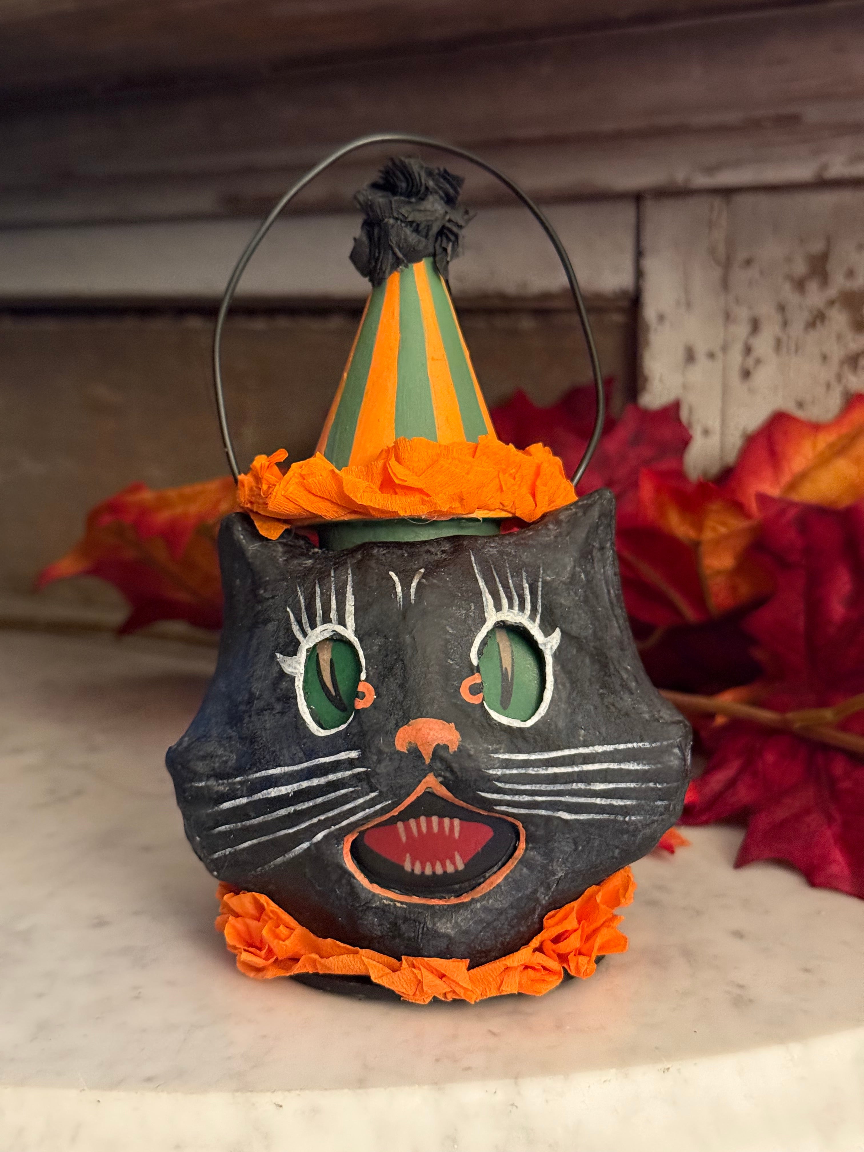 A black cat-shaped Halloween lantern with orange ruffled trim, green eyes, and a pointed orange and green striped hat, shown hanging against a rustic wooden and stone background.
