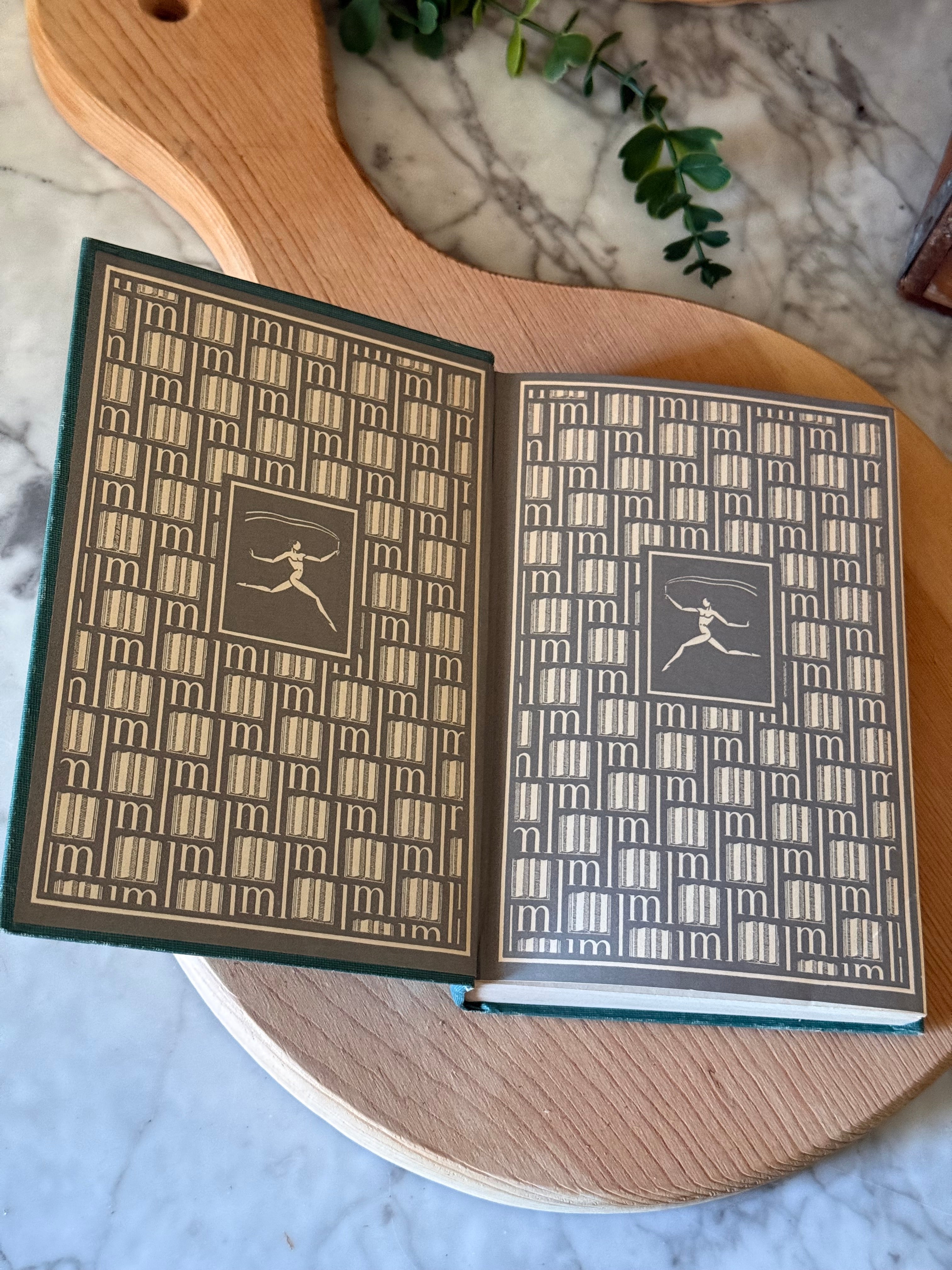 An open vintage hardcover book with patterned beige and dark green covers, displayed on a round wooden board over a marble surface.
