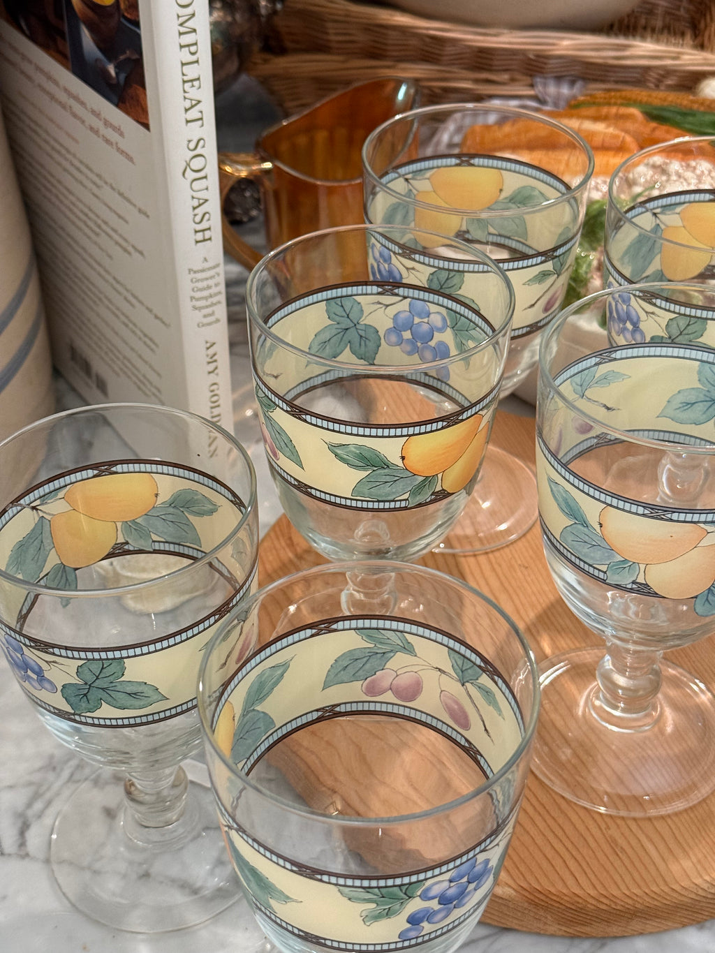 A set of clear glass stemware cups decorated with a vintage-style fruit pattern featuring lemons, blueberries, and green leaves on a cream background, arranged on a wooden board.