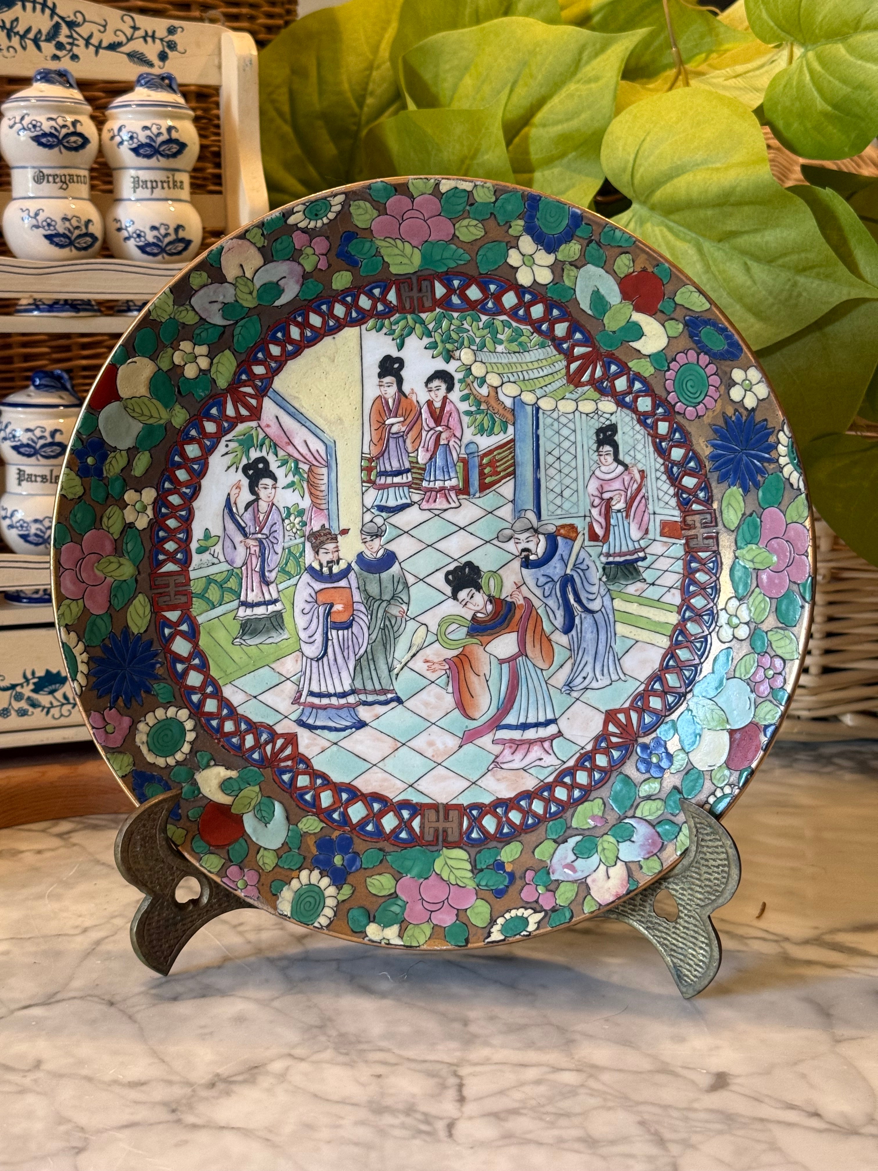 A round ceramic plate with a colorful hand-painted Chinese-style scene of women in a pavilion, displayed on a small stand on a marble surface with green leaves and ceramic jars in the background.