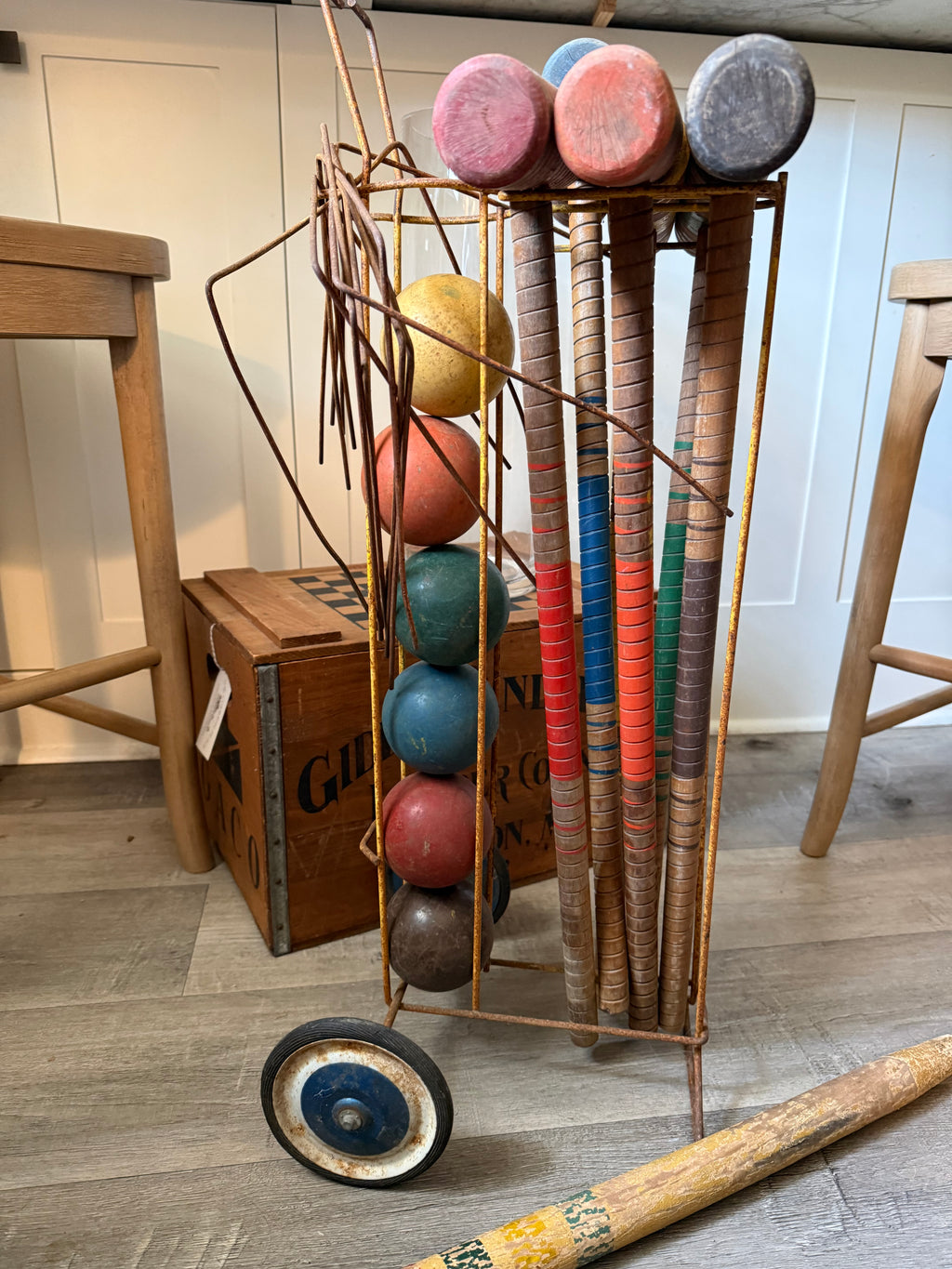 A vintage-style metal wheeled rack holding multiple cricket bats and round cricket balls, photographed indoors on a wood floor with stools and a crate in the background.