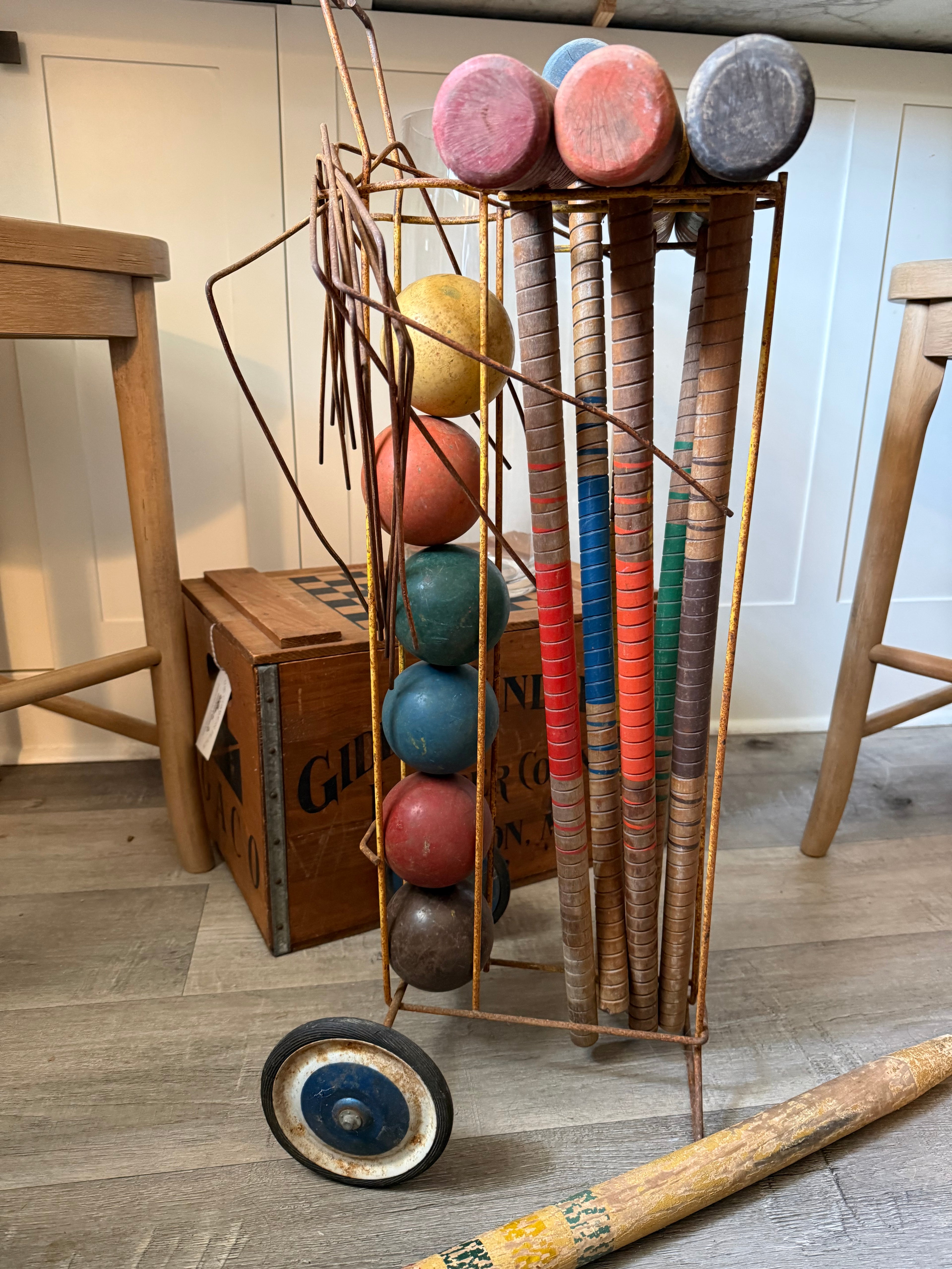 A vintage-style metal wheeled rack holding multiple cricket bats and round cricket balls, photographed indoors on a wood floor with stools and a crate in the background.