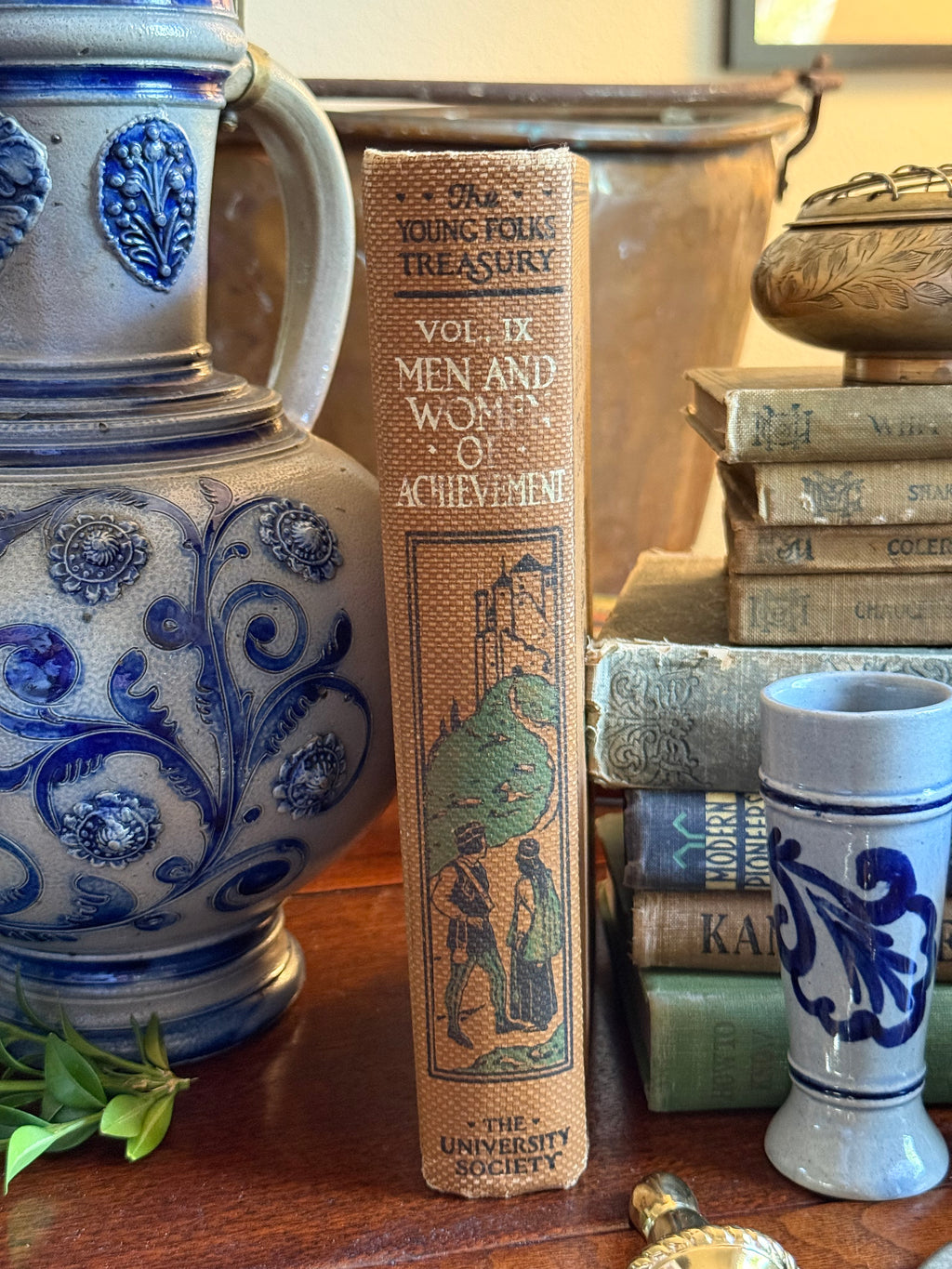 A vintage hardcover book titled 'One Young Folks Treasury Vol. IX Men and Wonders of Achievement' stands upright on a wooden table, surrounded by decorative ceramic vases, old books, and a small blue-and-white mug.