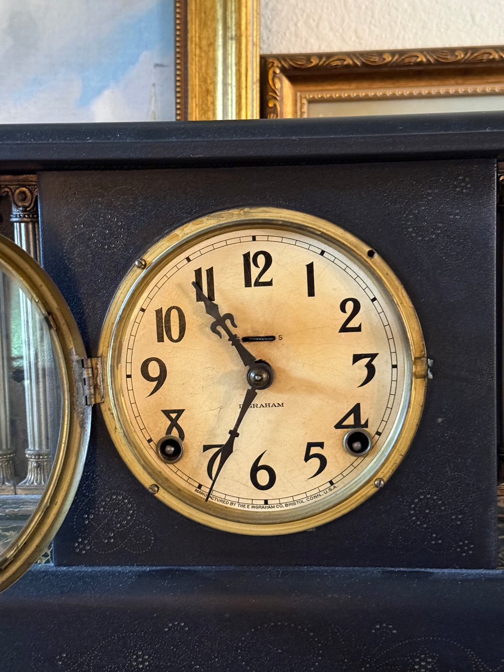 Close-up of an antique E. Ingraham Co. mantel clock's analog face. The cream-colored dial features elegant, vintage black Arabic numerals and ornate spade hands, framed by a polished brass bezel. The clock face is set against a dark black architectural casing with intricate textured patterns, with a glimpse of a gold picture frame and oil painting styled directly above it.