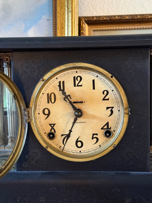 Close-up of an antique E. Ingraham Co. mantel clock's analog face. The cream-colored dial features elegant, vintage black Arabic numerals and ornate spade hands, framed by a polished brass bezel. The clock face is set against a dark black architectural casing with intricate textured patterns, with a glimpse of a gold picture frame and oil painting styled directly above it.