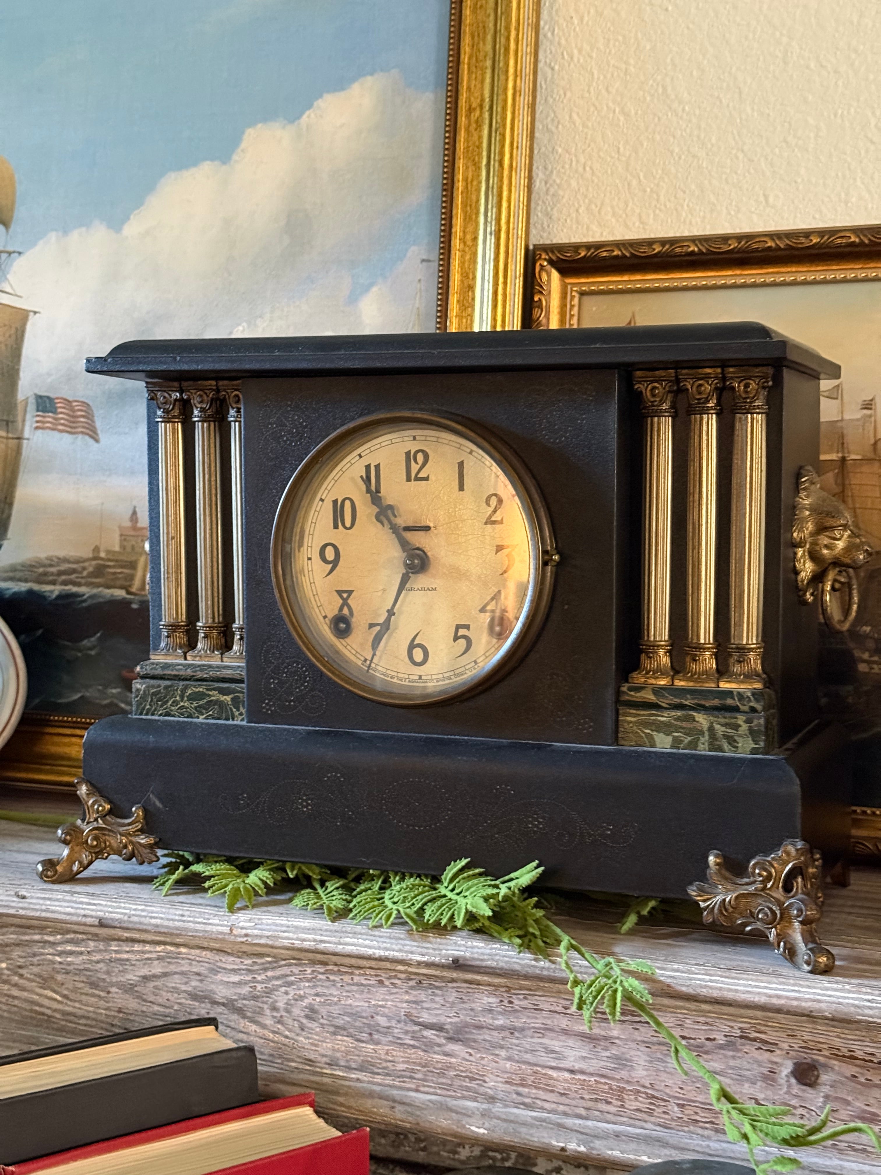 **`An antique E. Ingraham Co. mechanical mantel clock displayed in a home interior setting. The clock features a dark, black architectural hardwood casing accented with gold-toned metal pillars, elegant golden lion head details on the sides, and dark green faux-marble trim along its base and top columns. The round, cream-colored analog clock face displays clear Arabic numerals and ornate hands, styled next to a small green botanical garland on a wooden shelf.`**