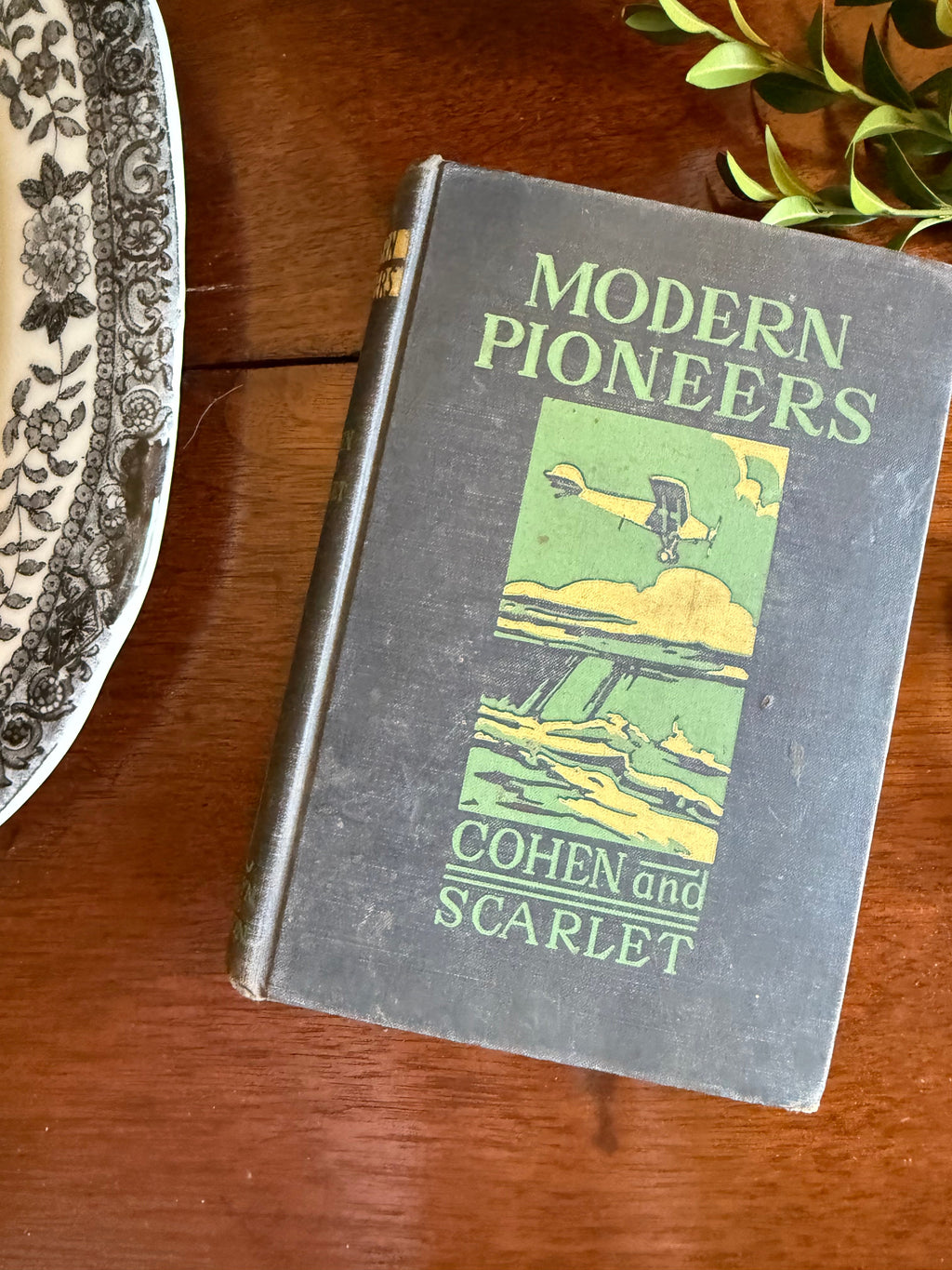 A vintage hardcover book titled 'Modern Pioneers' by Cohen and Scarlet, featuring a dark gray cover with green and yellow illustration and text, resting on a wooden table.