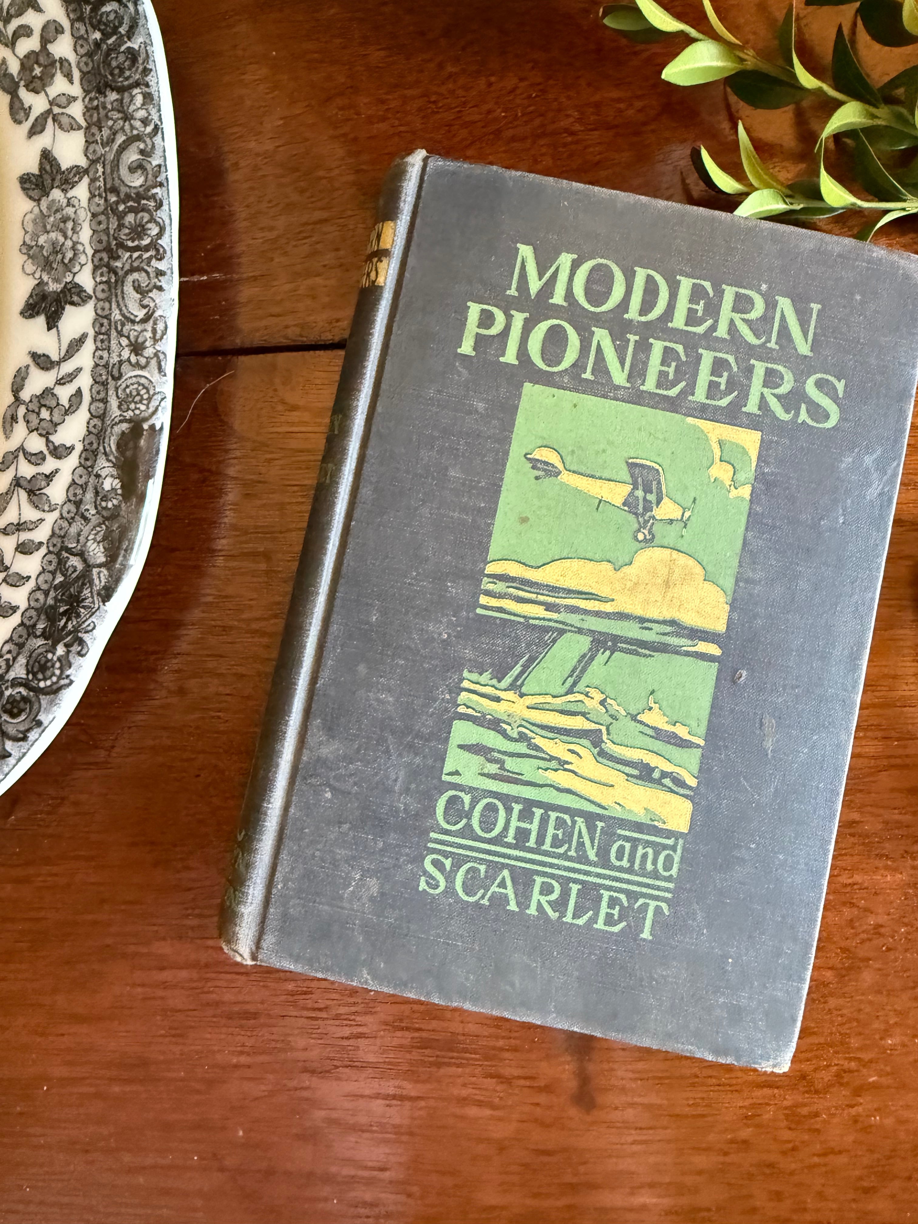A vintage hardcover book titled 'Modern Pioneers' by Cohen and Scarlet, featuring a dark gray cover with green and yellow illustration and text, resting on a wooden table.