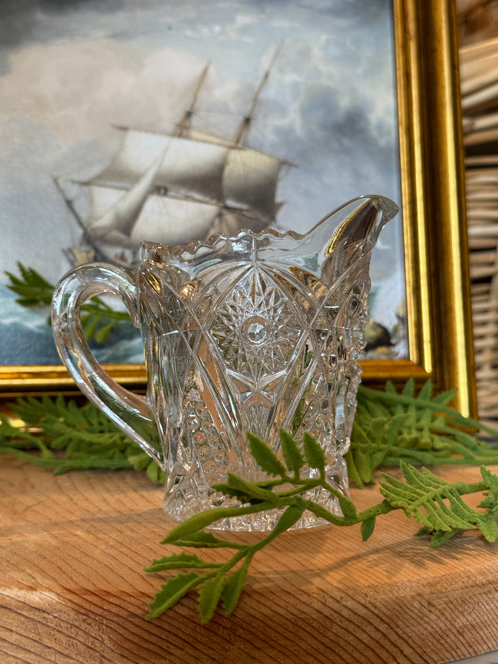 A clear cut-crystal glass pitcher with a handle and spout, displayed on a wooden surface next to green foliage, with a framed ship painting in the background.