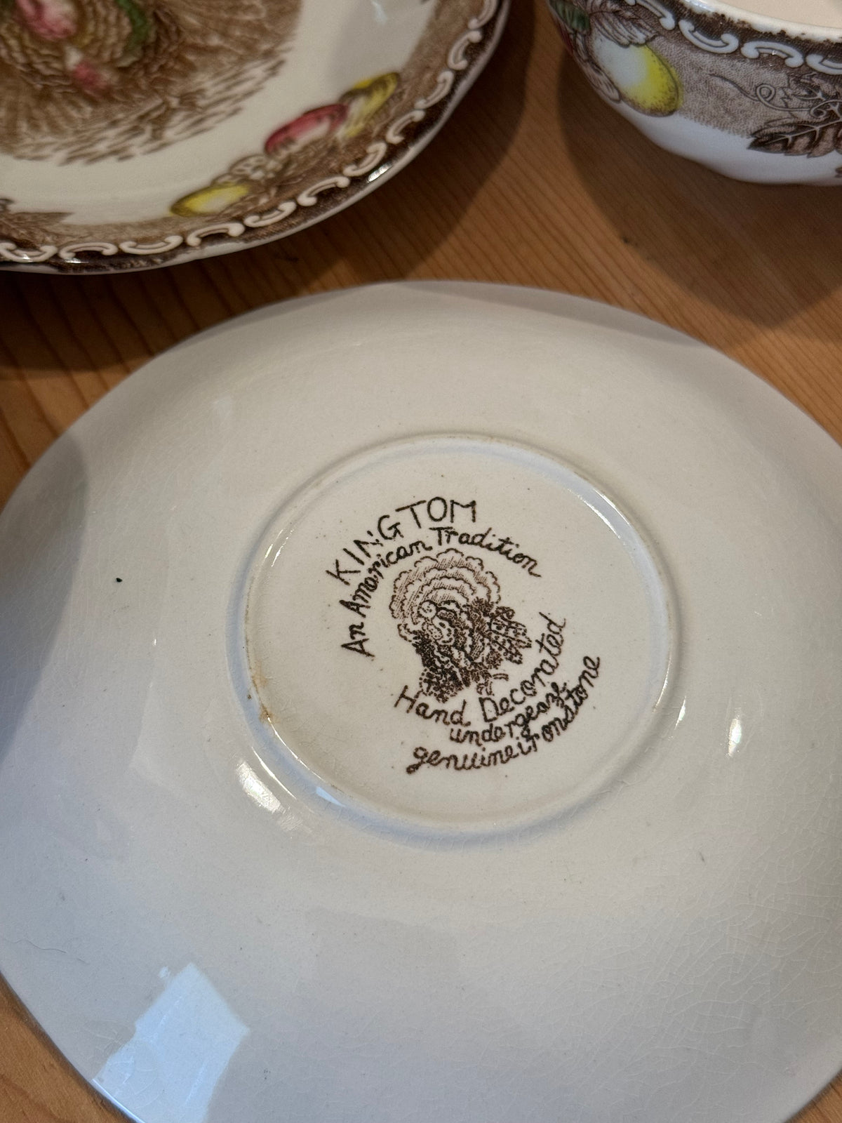 Close-up of the bottom of a white ceramic plate or saucer showing printed branding text that reads 'KIN TOM with a floral emblem and smaller decorative text, placed on a wooden surface with other patterned dishes partially visible.