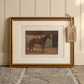 A framed vintage print of a brown horse, shown leaning against a cream paneled wall on a stone floor. The print has a white mat and is enclosed in a gold frame, with a hanging paper tag that reads 'Captain Lewis'.