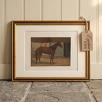 A framed vintage print of a brown horse, shown leaning against a cream paneled wall on a stone floor. The print has a white mat and is enclosed in a gold frame, with a hanging paper tag that reads 'Captain Lewis'.