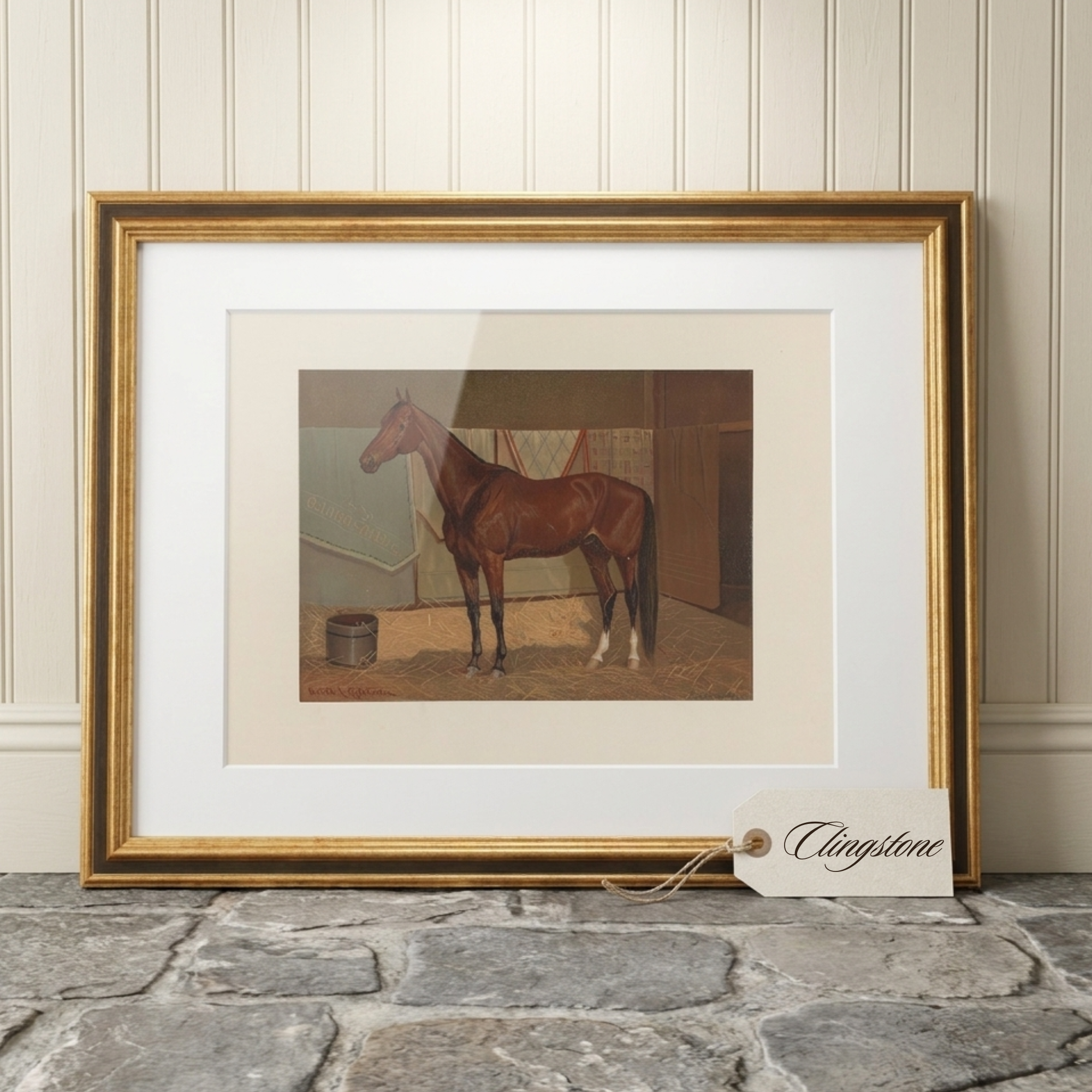 A framed art print of a brown horse in a stable, styled on a stone floor against a cream paneled wall. The print features a white mat and a wide gold frame, with a hanging tag reading 'Clingstone'.