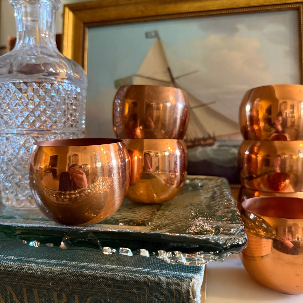 Decorative vintage bar cart with copper cups on a surface with books and a framed picture in the background.