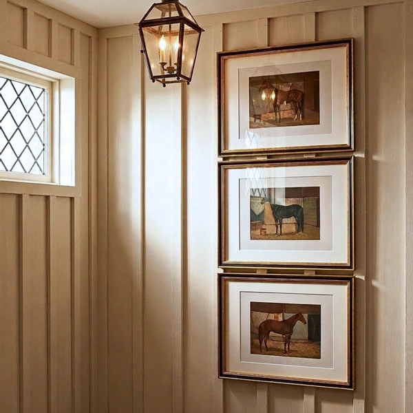 Set of three framed vintage racehorse art prints stacked vertically on a warm wood panel wall in an entryway with a gas lantern