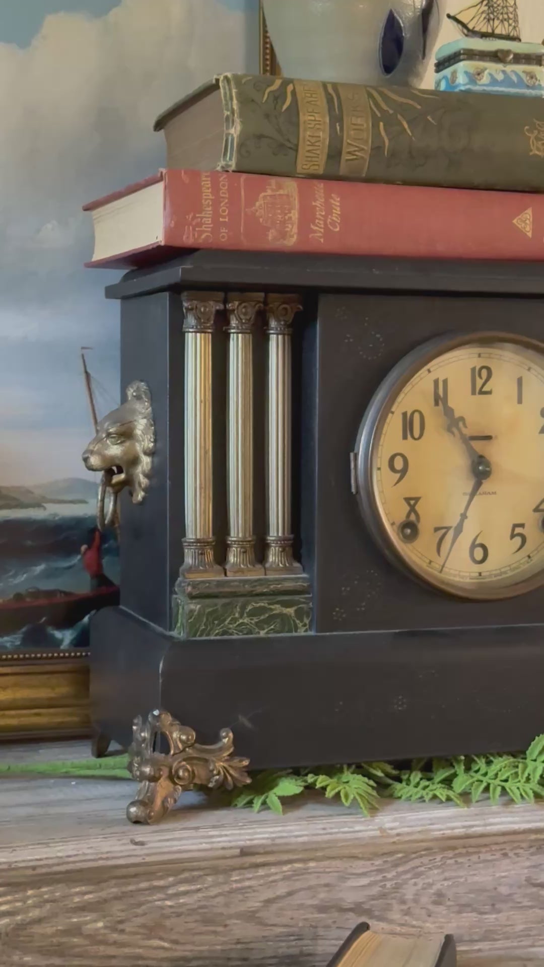 Video of an antique E. Ingraham Co. mechanical mantel clock showcasing its historic details. The camera slowly pans and focuses on the front facade of the dark hardwood architectural casing, adorned with elegant golden lion head accents on the sides, faux-marble green trim bases, and classical columns framing the clock face. The cream-colored analog dial features vintage numerals and ornate hands, captured in warm, natural light as a stylized staging piece.