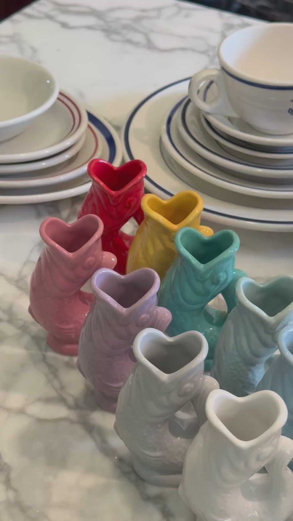 A panning video showcasing a colorful collection of mini ceramic Gluggle Jugs in shades of pink, red, yellow, lilac, teal, and white, arranged on a marble surface.