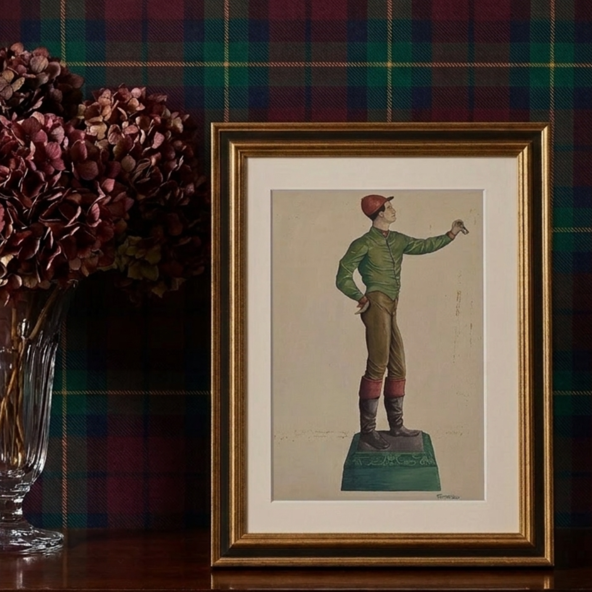 A framed vintage illustration of a jockey standing on a pedestal, displayed on a dark wooden surface against a green and red plaid wall, with a glass vase of dried burgundy flowers to the left.