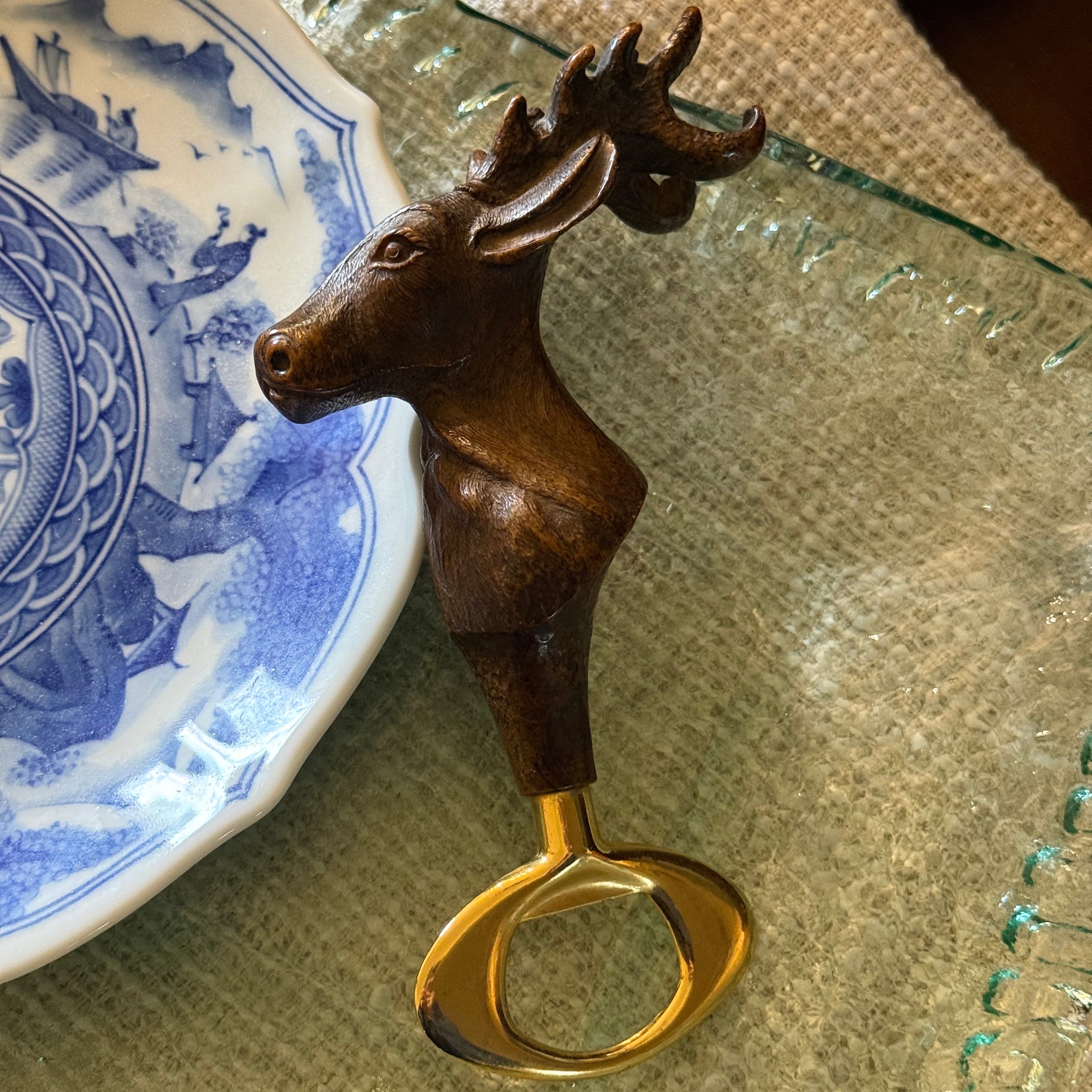 Close-up of the vintage stag bottle opener next to a blue patterned plate, showcasing its scale and "hunting lodge" style.