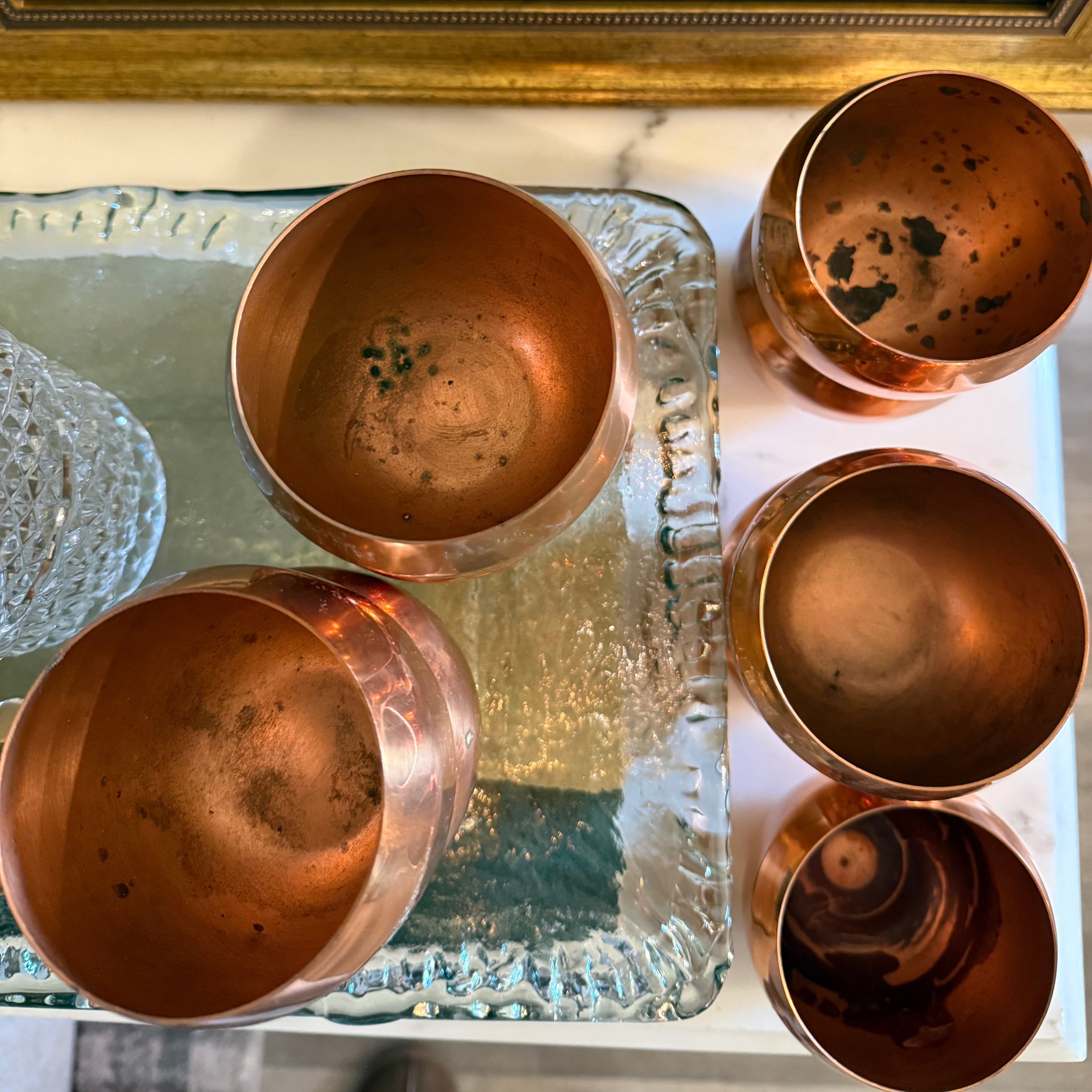 Set of five copper cups on a glass tray with a framed picture of a sailboat in the background.