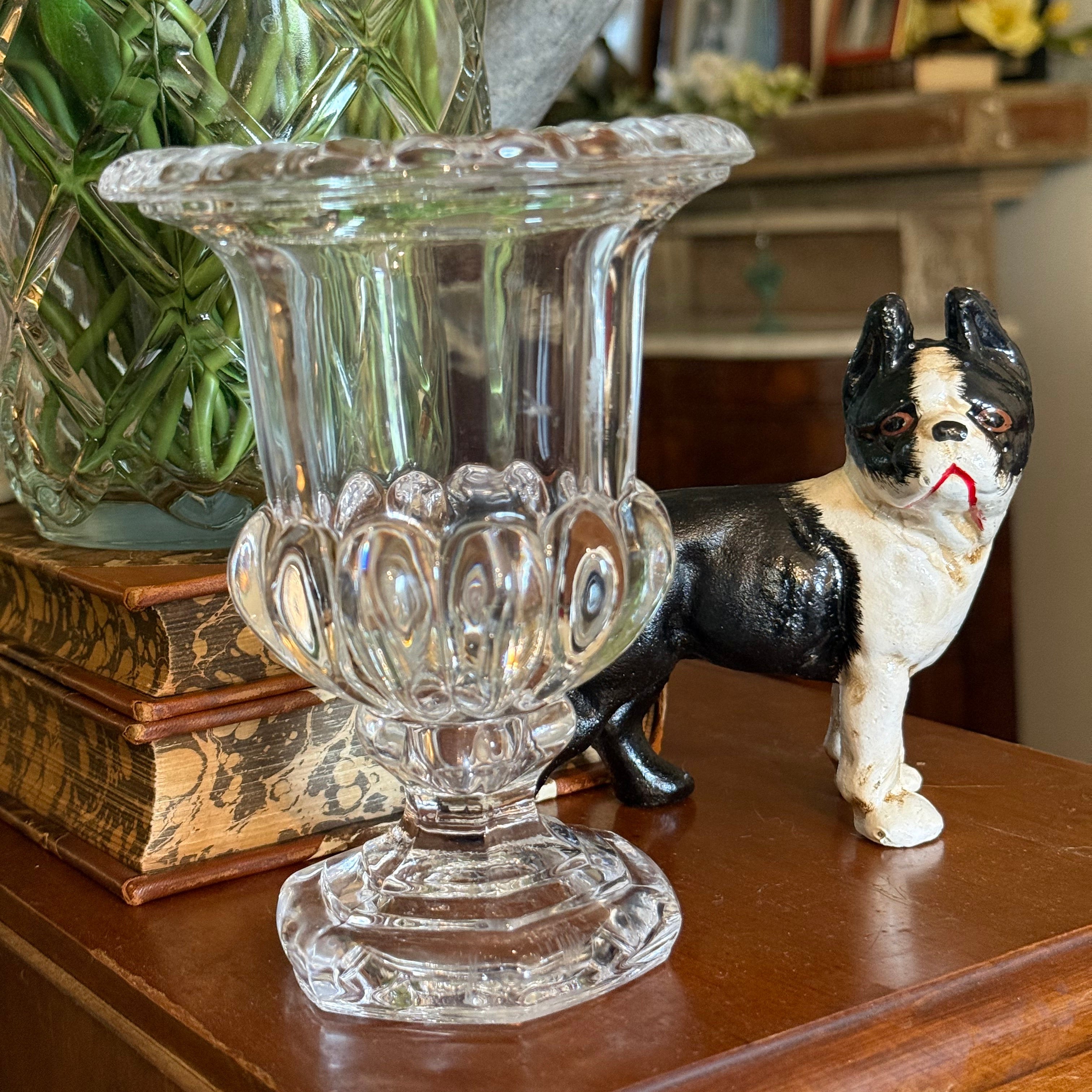 Front view of a clear vintage mini crystal urn vase with a footed pedestal base, standing next to a decorative dog figurine.